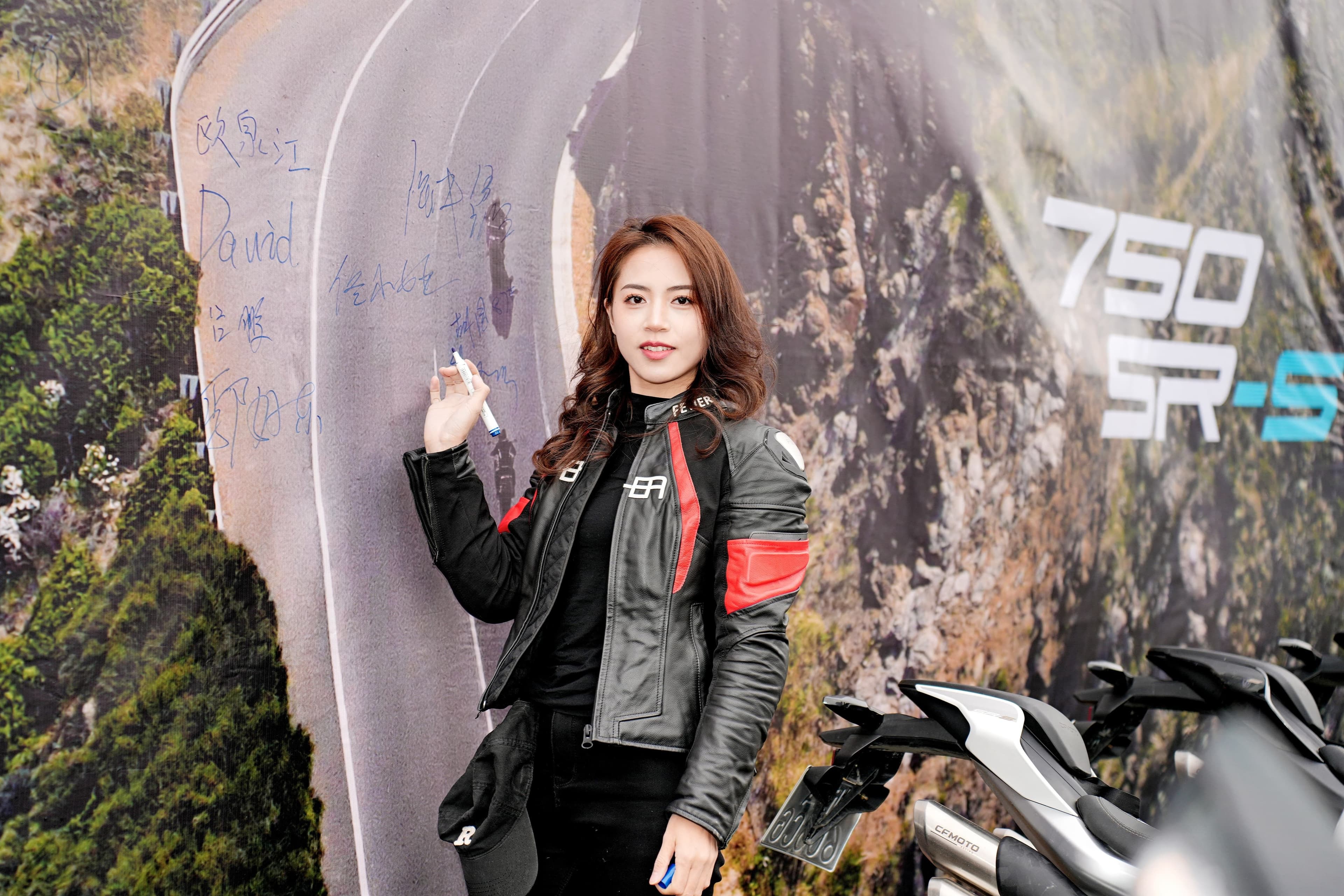 Woman in black and red motorcycle jacket posing by "750 SR-S" brand backdrop.